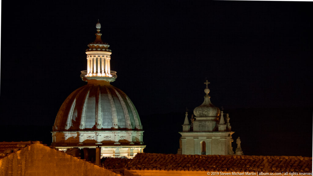 Large illuminated ribbed church dome with columned lantern above dark tiled roofs, accompanied by a smaller domed structure.