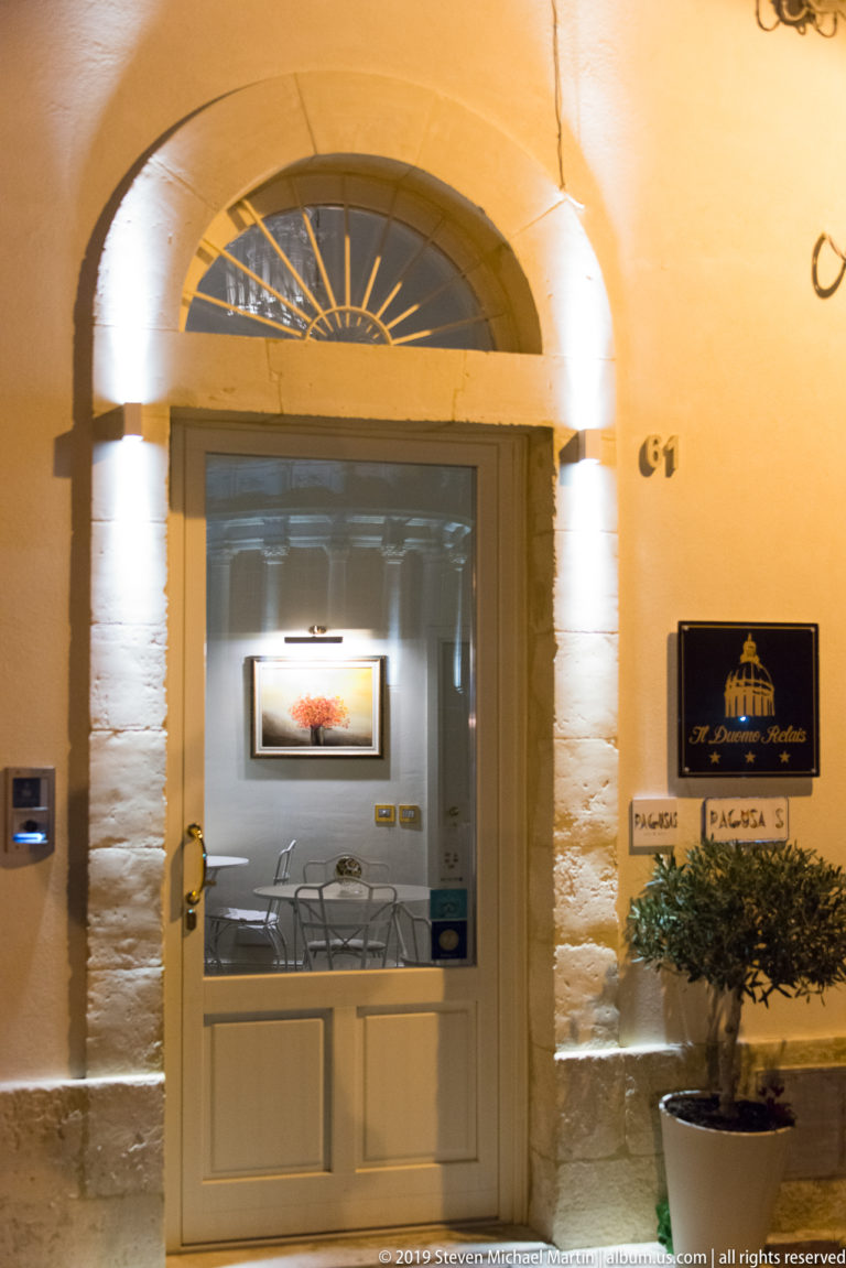 Illuminated arched entrance with stone jambs, glazed door, fanlight, Ragusa plaques, and a small seating area visible inside.
