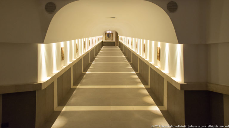 Broad white vault opening onto a long symmetrical corridor with illuminated wall displays, side ledges, and a distant doorway.