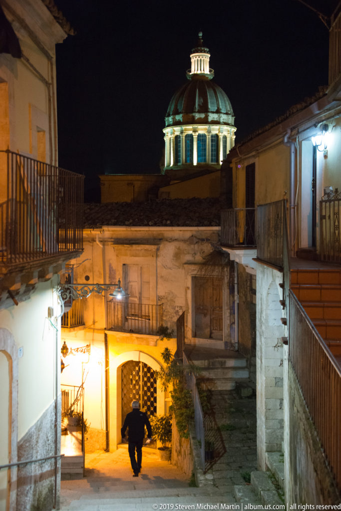 Illuminated church dome above weathered rooftops and balconies, with a lone pedestrian walking through a lit passage below.