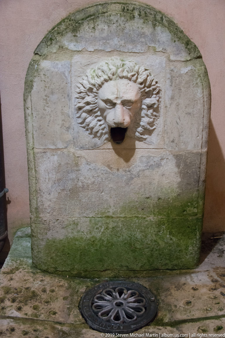 Weathered stone wall fountain with a carved lion-mask spout, green staining at the base, and a round drain cover below.