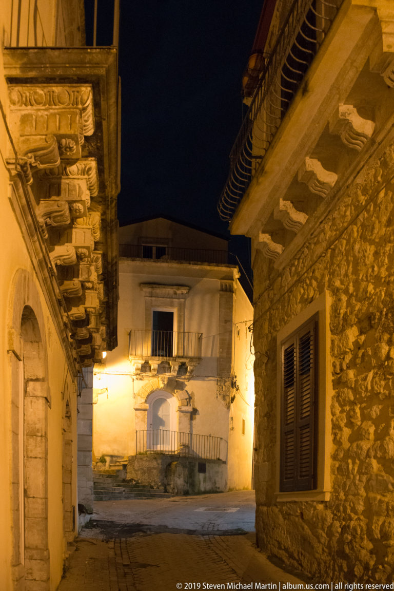 Night passage between historic buildings, with rough stone wall, shuttered window, carved doorway, balconies, and deep projecting eaves.