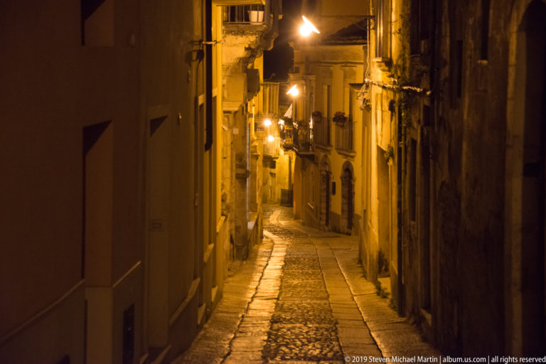 Narrow stone-paved lane descending between old buildings under a line of warm streetlamps at night.