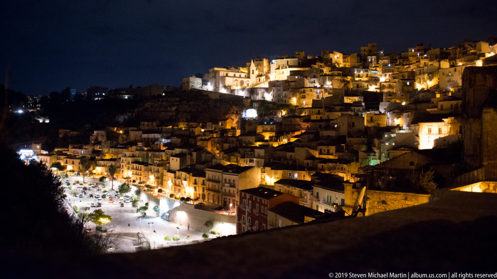 Night panorama of Ragusa climbing a steep hillside, with illuminated houses, streets, and a bright open square below.