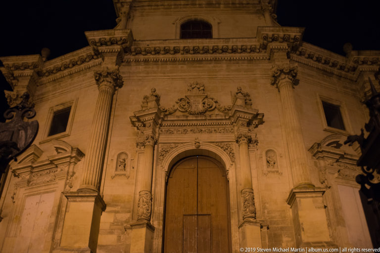 Upward nighttime view of a Baroque church entrance with tall wooden doors, carved stone portal, columns, niches, and ornate cornices.