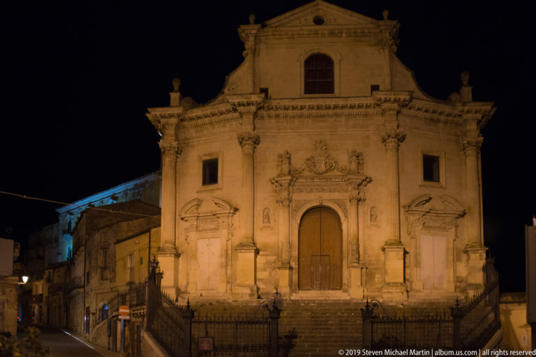 Frontal floodlit Baroque church façade with three doorways, carved crest, pilasters, broad steps, and iron railings at night.
