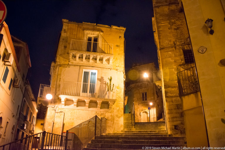 Stone steps rising between warmly lit residential buildings with balconies, railings, windows, and street lamps at night.