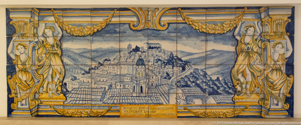 Large ceramic tile panel showing a hillside cityscape of Ragusa with a domed church, framed by painted figures, garlands, and scrollwork.