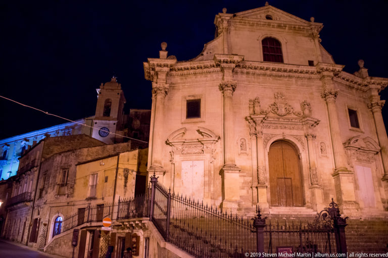 Floodlit Baroque church façade with three portals, carved stonework, steps, iron railings, and adjoining buildings under a dark sky.