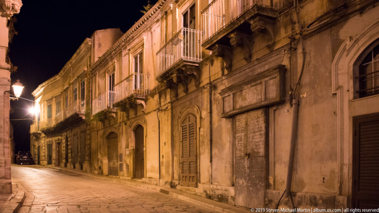 Oblique nighttime view along stone townhouses with shuttered doors, carved balcony brackets, metal railings, and warm street lighting.