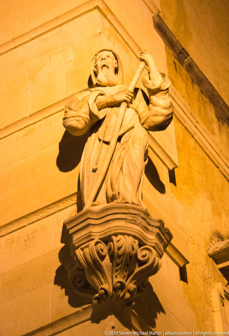 Carved stone statue of a robed saint holding a long staff, mounted on an ornate bracket against a lit church wall.