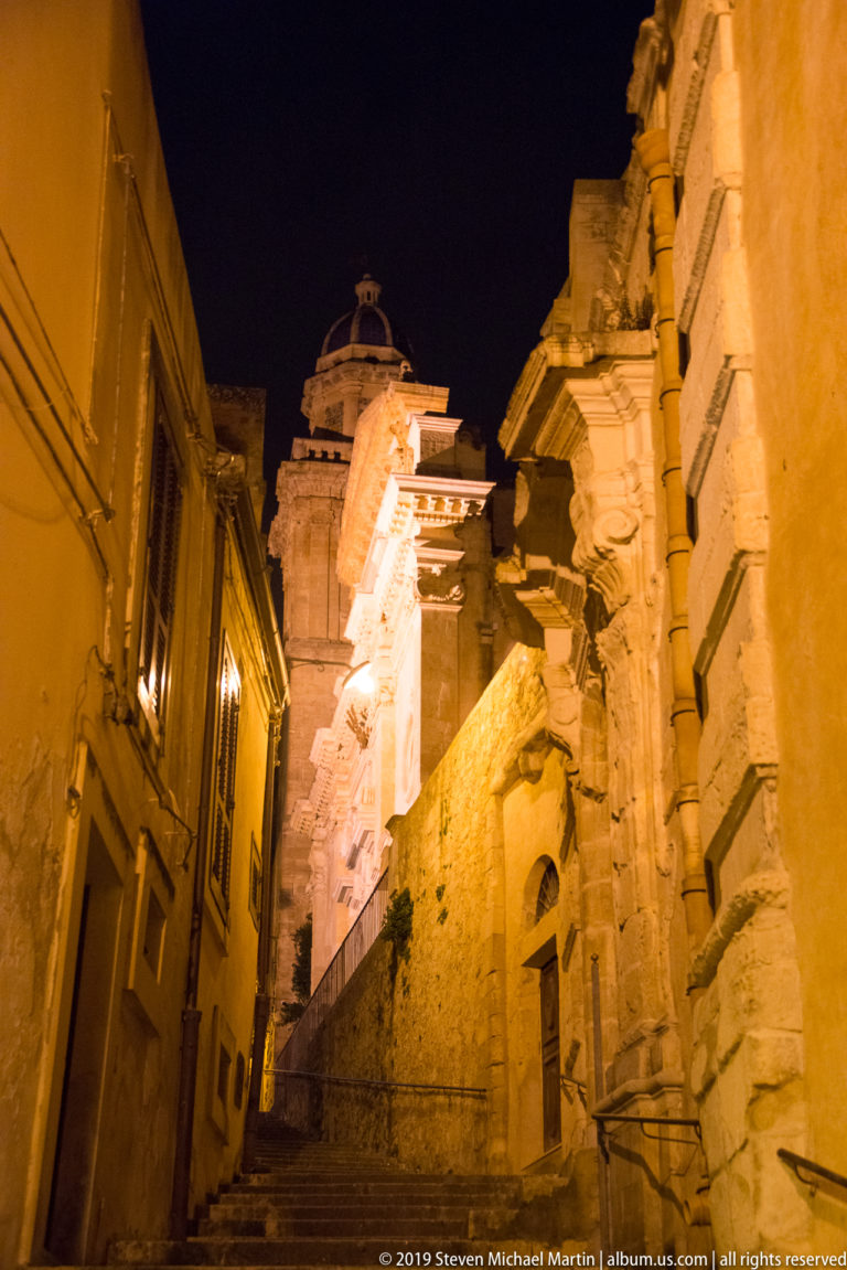 Narrow stone staircase climbing between tall buildings toward a brightly illuminated ornate façade under a dark night sky.