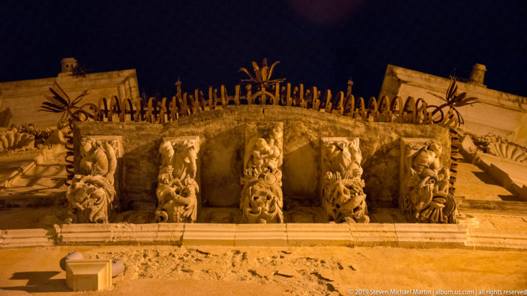 Five carved stone grotesque corbels beneath a balcony, each with a different expressive figure, viewed from below at night.