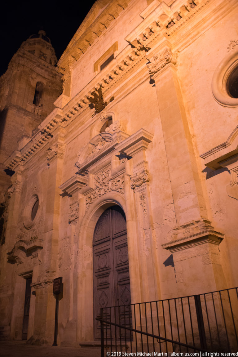 Illuminated stone church wall with ornate arched wooden doorway, pilasters, circular opening, cornices, and a low street railing.