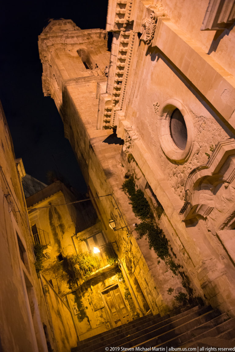 Steep upward view of a stone church wall with circular oculus, carved cornices, and a narrow neighboring building at night.