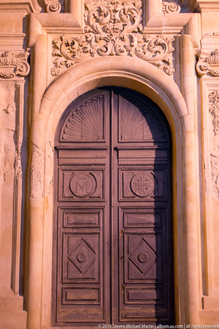 Tall paneled wooden double door set within an ornate carved stone arch with scrollwork and molded Baroque decoration.