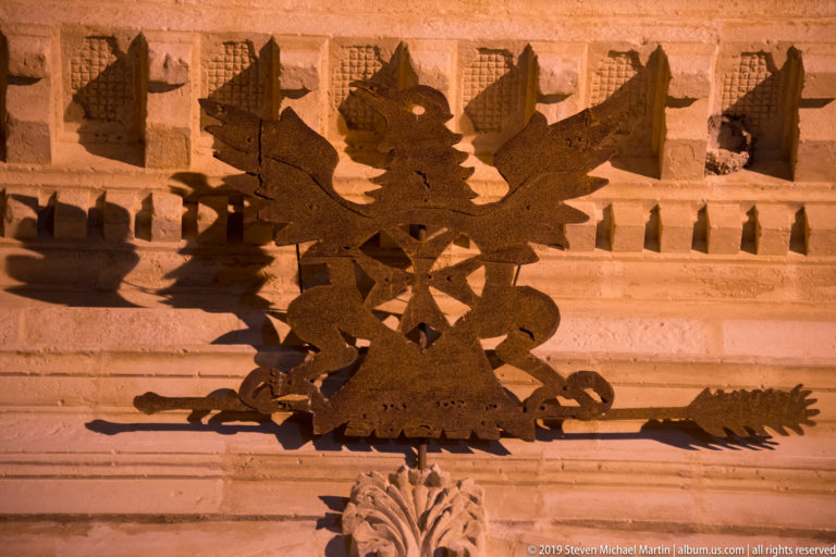 Dark symmetrical wrought-iron crest with scrolling forms casting shadows across a warmly lit carved stone façade.