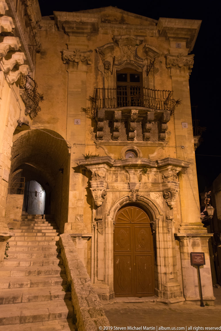 Ornate stone doorway with carved surround and balcony beside a vaulted passage containing a steep flight of steps.