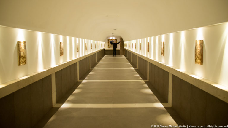 Long symmetrical corridor lined with small wall artworks and repeated vertical lights, with a person standing near the distant end.