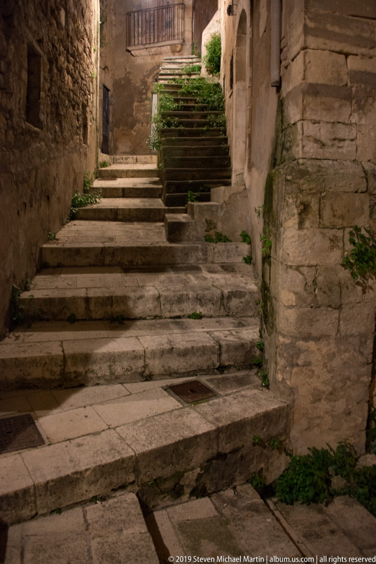 Worn stone steps rising through a narrow alley with stacked planters, rough masonry walls, arched openings, and small plants.