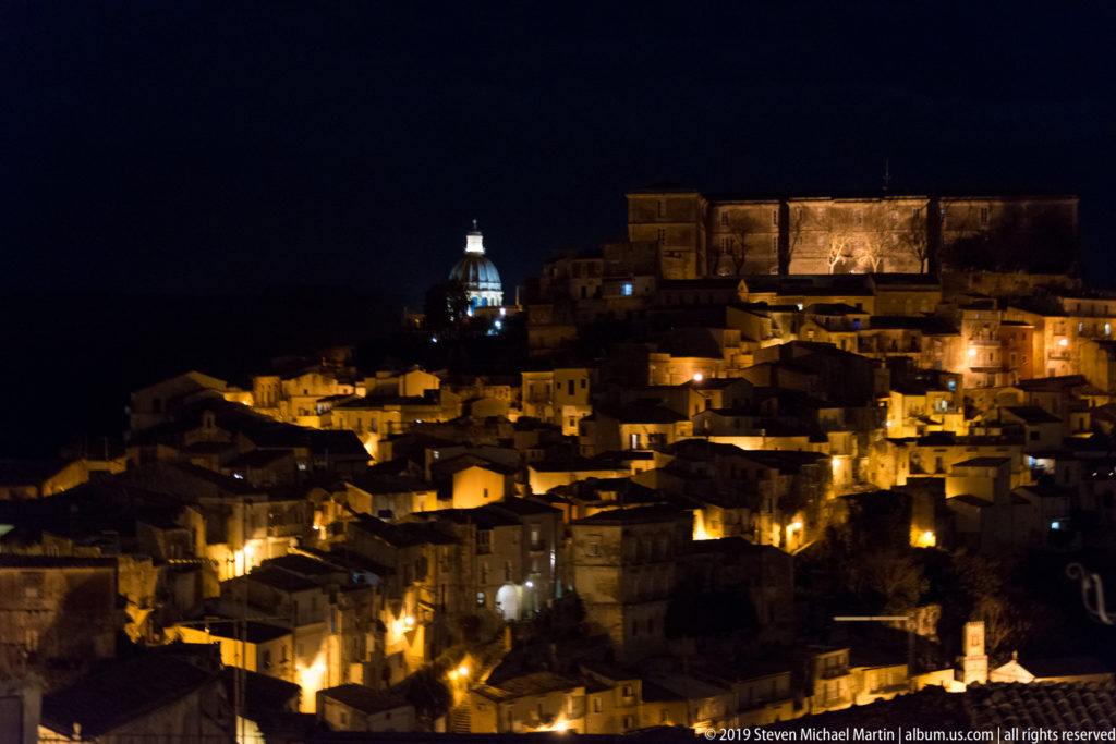 Tight night view of Ragusa Ibla with glowing houses, a bright domed tower, and a large complex on the upper ridge.