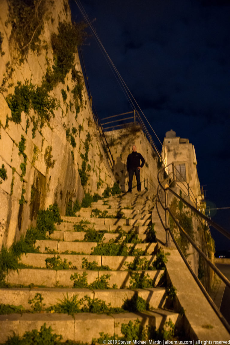 Steep worn stone steps between masonry walls with iron handrail, overhead wires, vegetation, and a person climbing near the top.