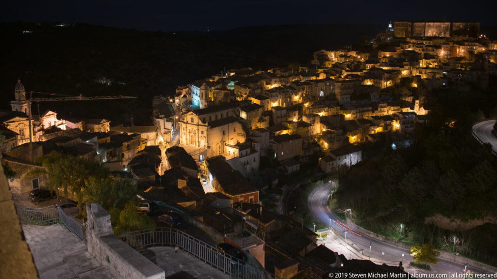 Elevated nighttime panorama of Ragusa Ibla with illuminated hillside houses, winding roads, and a prominent church complex below.