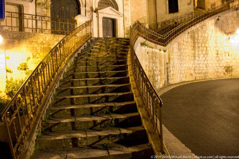 Broad worn stone staircase with curving iron railings rising to a doorway beside a bending road at night.