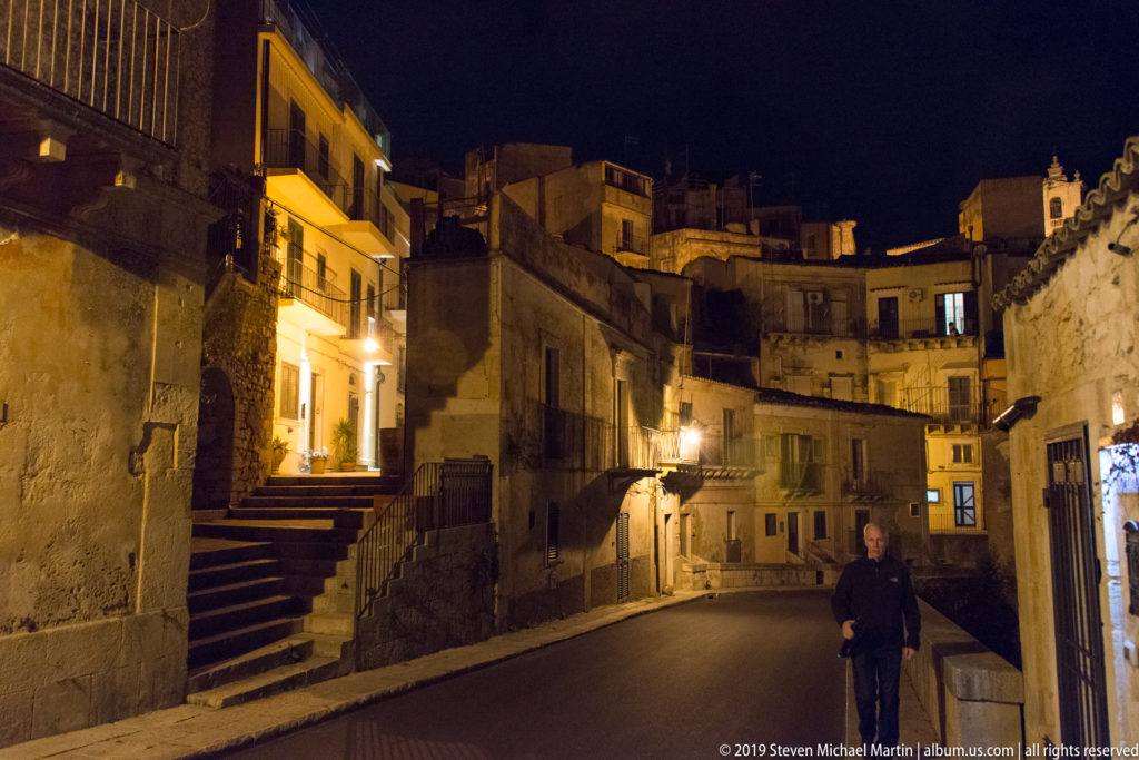 Steep residential lane with stone steps, closely packed houses, warm streetlights, balconies, and a lone pedestrian at night.
