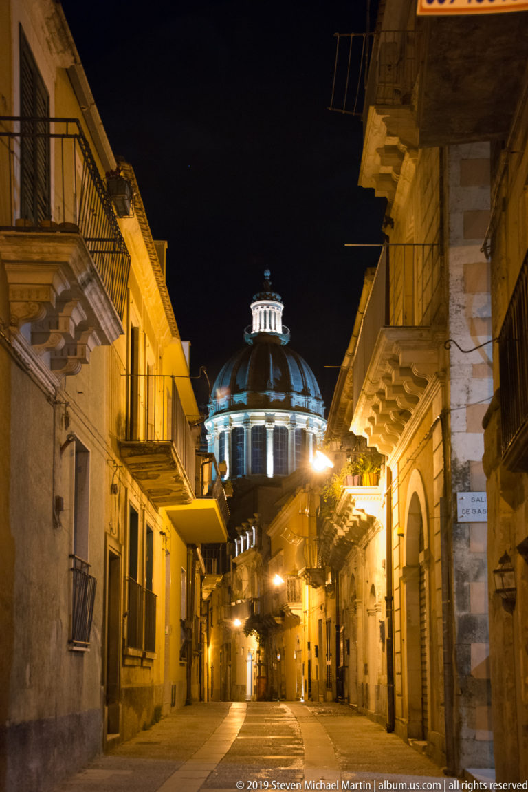 Narrow nighttime street lined with balconies and stone façades, ending in a brightly illuminated dome.