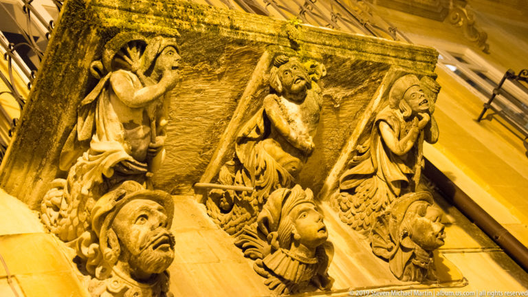 Upward close view of carved stone grotesque balcony supports with expressive faces and deep shadows under warm lighting.