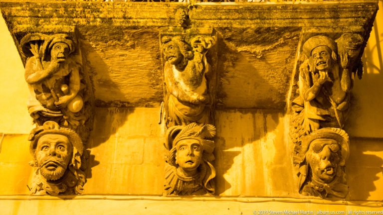 Close view of carved stone balcony supports with expressive human faces and grotesque figures under warm night lighting.