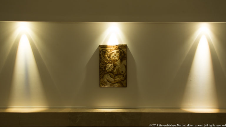Small carved rectangular relief centered on a pale wall beneath focused spotlights in a modern interior corridor.