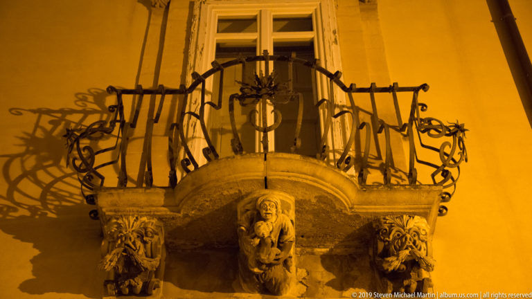 Curved wrought-iron balcony supported by carved stone grotesque masks and brackets, casting strong shadows on a yellow-lit wall.