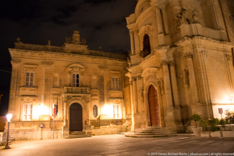 Illuminated palazzo with balcony beside the projecting façade of a large Baroque church in a quiet square.