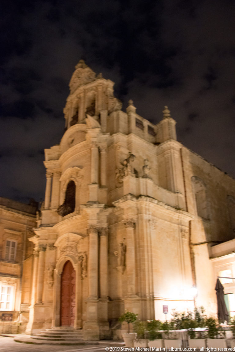 Oblique nighttime view of a large Baroque church with layered columns, niches, carved stonework, and a tall central façade.