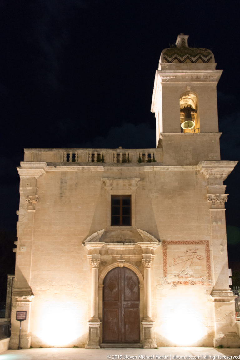 Illuminated stone church façade with wooden portal, shallow pediment, weathered wall surfaces, and a square bell turret.