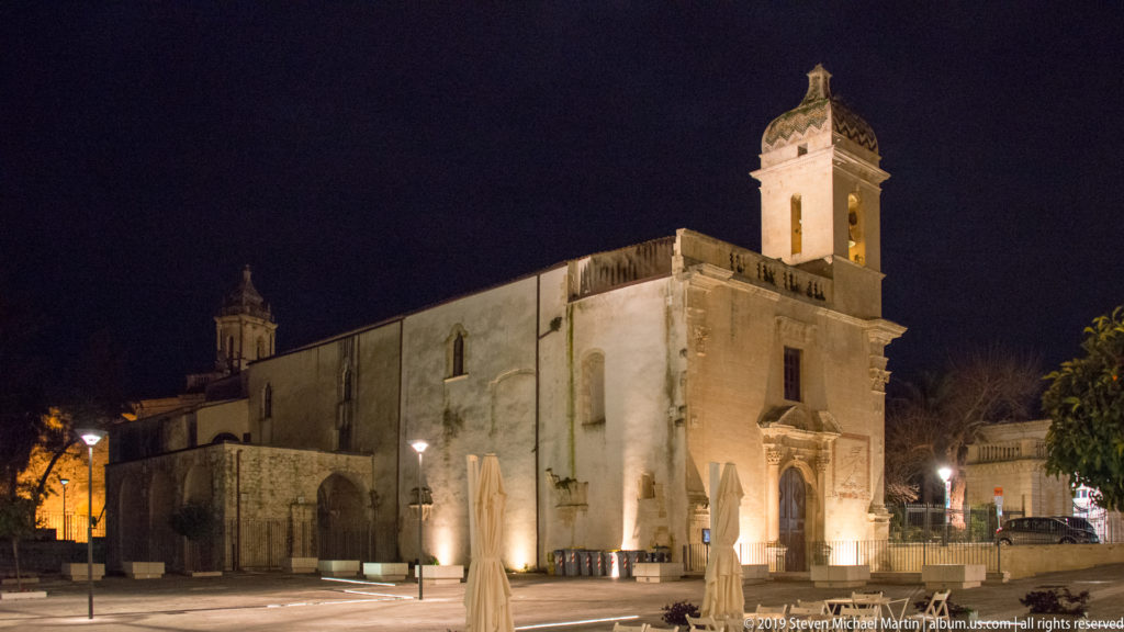 Floodlit stone church with square bell tower beside a broad, nearly empty paved square at night.