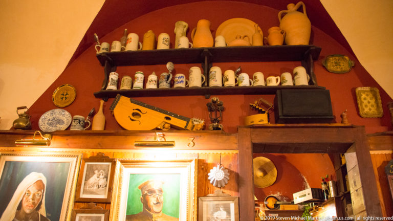 Restaurant wall display with ceramic vessels, framed portraits, a stringed instrument, shelves, and dark wood furniture.