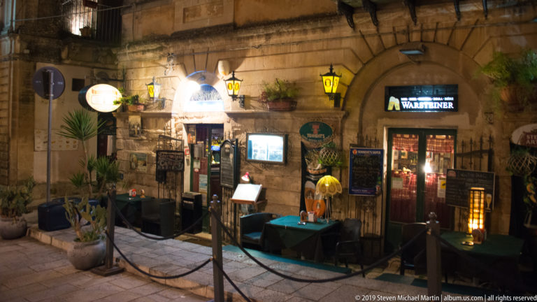 Restaurant entrance beneath stone arches with signs, lanterns, plants, small tables, and a roped outdoor seating area.