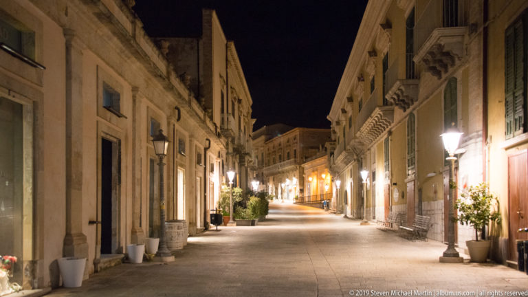 Quiet stone street at night lined with balconies, wall lamps, potted plants, and closely spaced historic buildings.