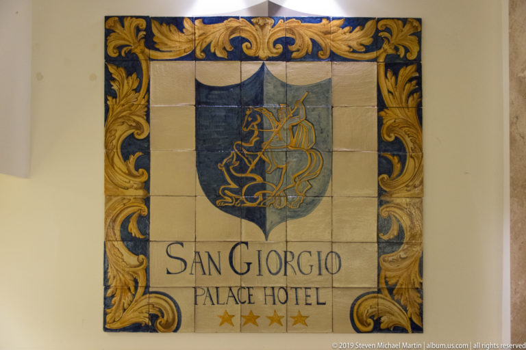 Ceramic tile sign reading “San Giorgio Palace Hotel,” with a blue shield, stylized sailing ship, ornate gold border, and four stars.