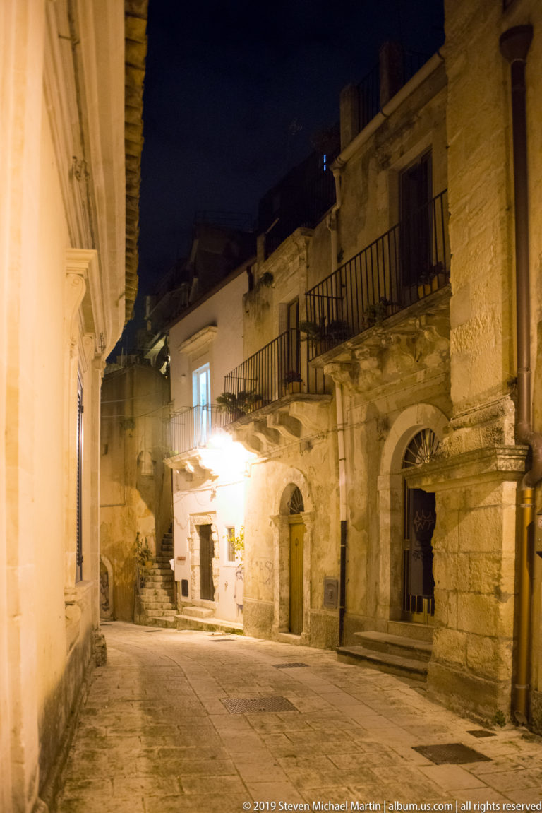 Empty stone-paved street curving between historic masonry buildings with arched doorways, iron balconies, and warm exterior lights.