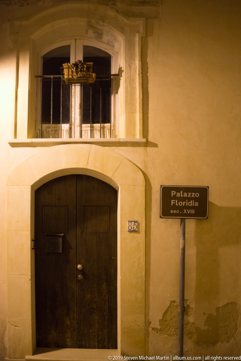 Arched wooden doorway in a pale façade beside a sign reading “Palazzo Floridia 1600–XVIII,” with a window above.