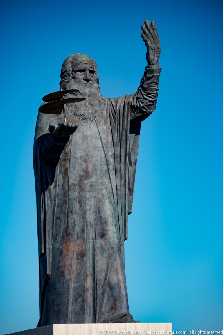 Bronze Leonardo da Vinci holding a winged flying-machine model in one hand and raising the other against blue sky.