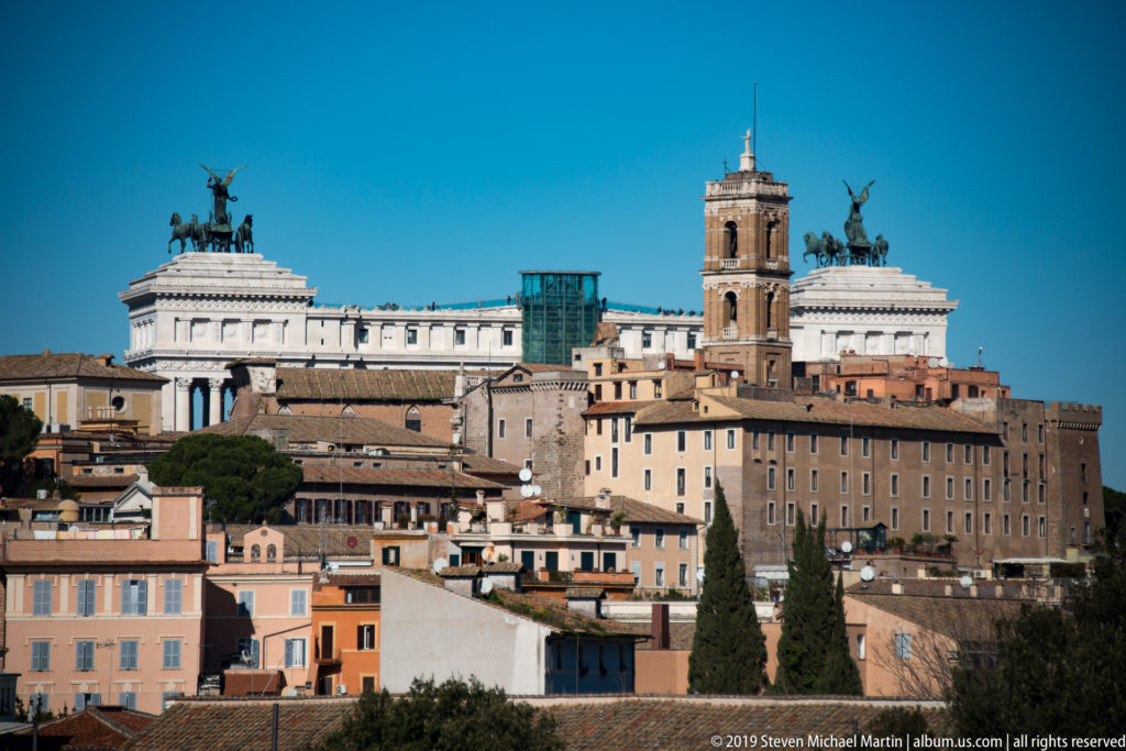 Rome skyline with the white Vittoriano, bronze quadrigas, Palazzo Senatorio clock tower, and dense historic buildings.