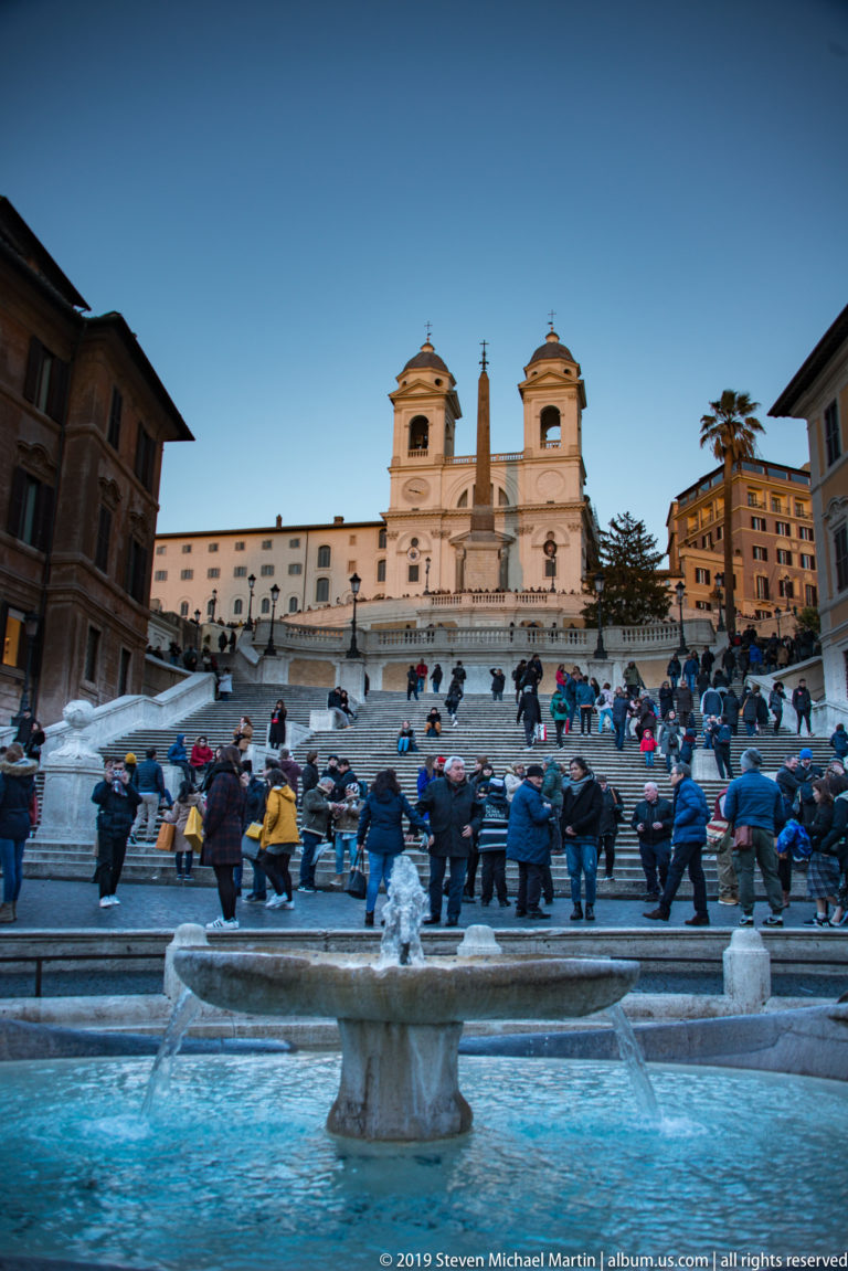 Spanish Steps at dusk with seated crowds, Barcaccia Fountain below, and Trinità dei Monti against a blue sky.