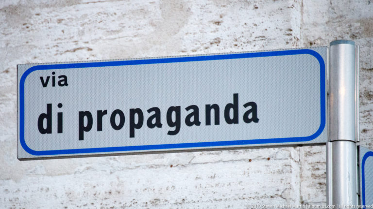 Blue-bordered street sign reading “via di propaganda” mounted on a pale stone wall in Rome.