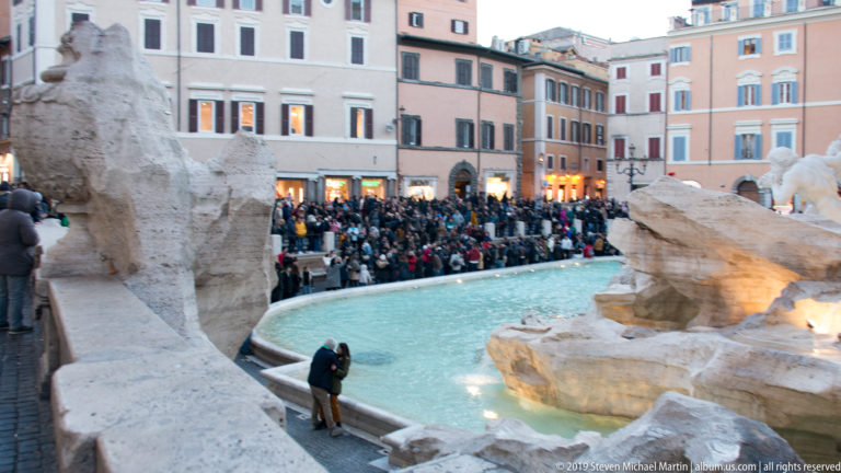 Turquoise Trevi Fountain basin and pale rockwork with a dense crowd gathered in the piazza beyond.