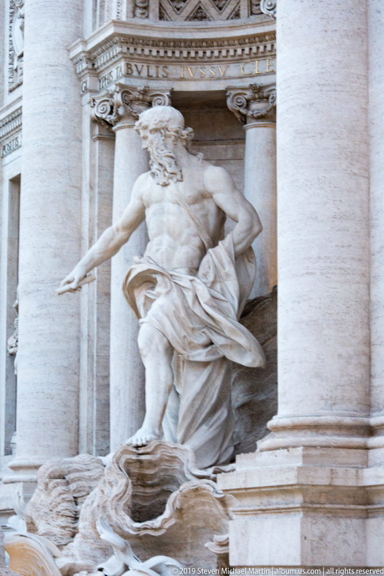Side view of the bearded Oceanus statue extending one arm within the central bay of the Trevi Fountain.